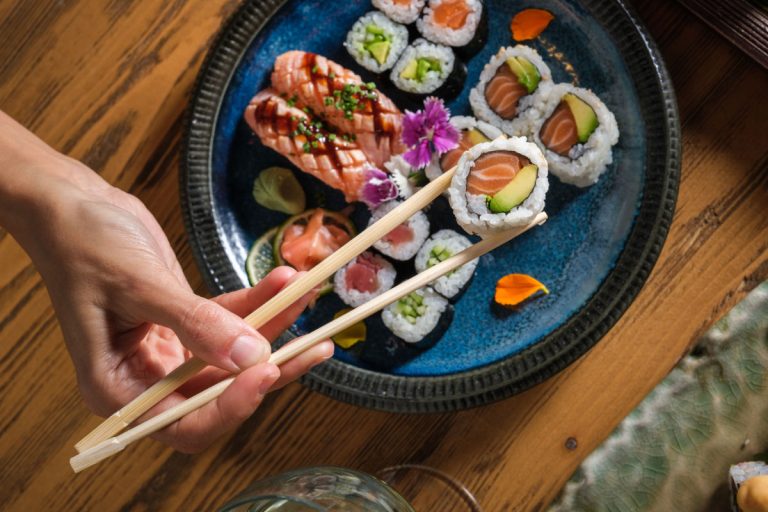 Top view of crop anonymous person with bamboo chopsticks taking tasty sushi roll with raw salmon and avocado from set